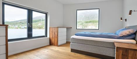 A bedroom with a large window, a bed, a wooden dresser, and a view of a landscape.