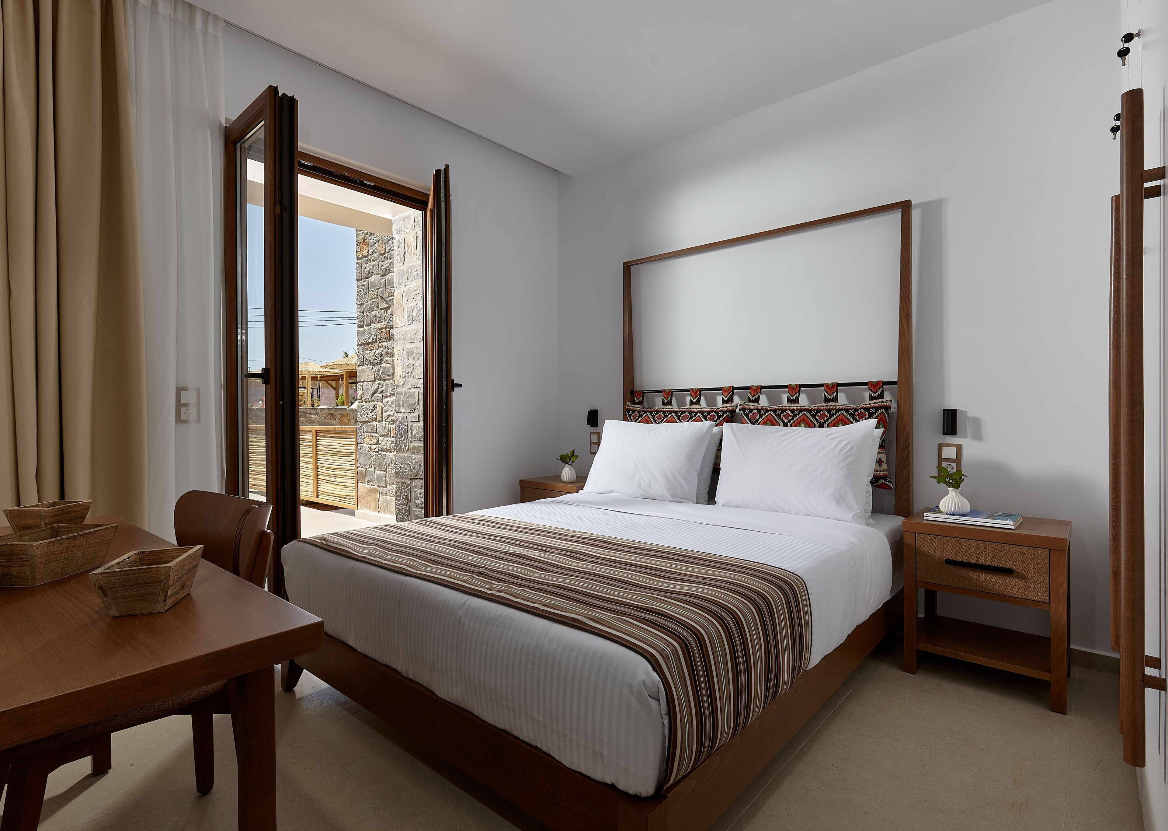 A bedroom with a bed, bedside tables, a wooden nightstand, a table with chairs, and a view of the outside.