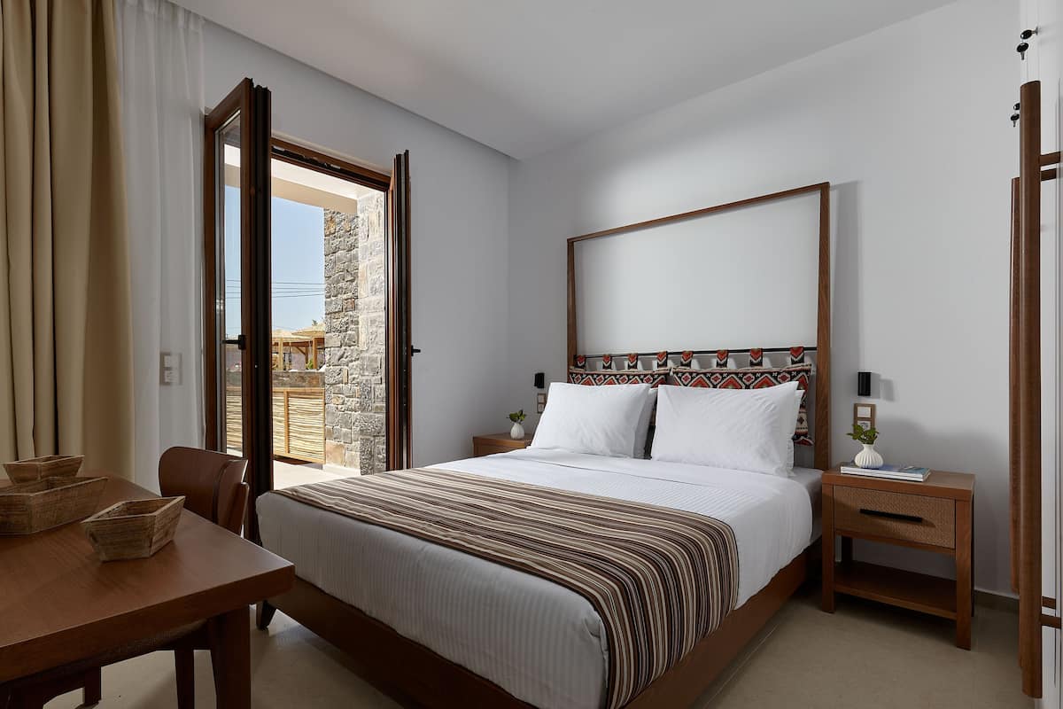 Ein Schlafzimmer mit einem Bett, Nachttischen, einem Holznachtkästchen, einem Tisch mit Stühlen und Blick ins Freie.