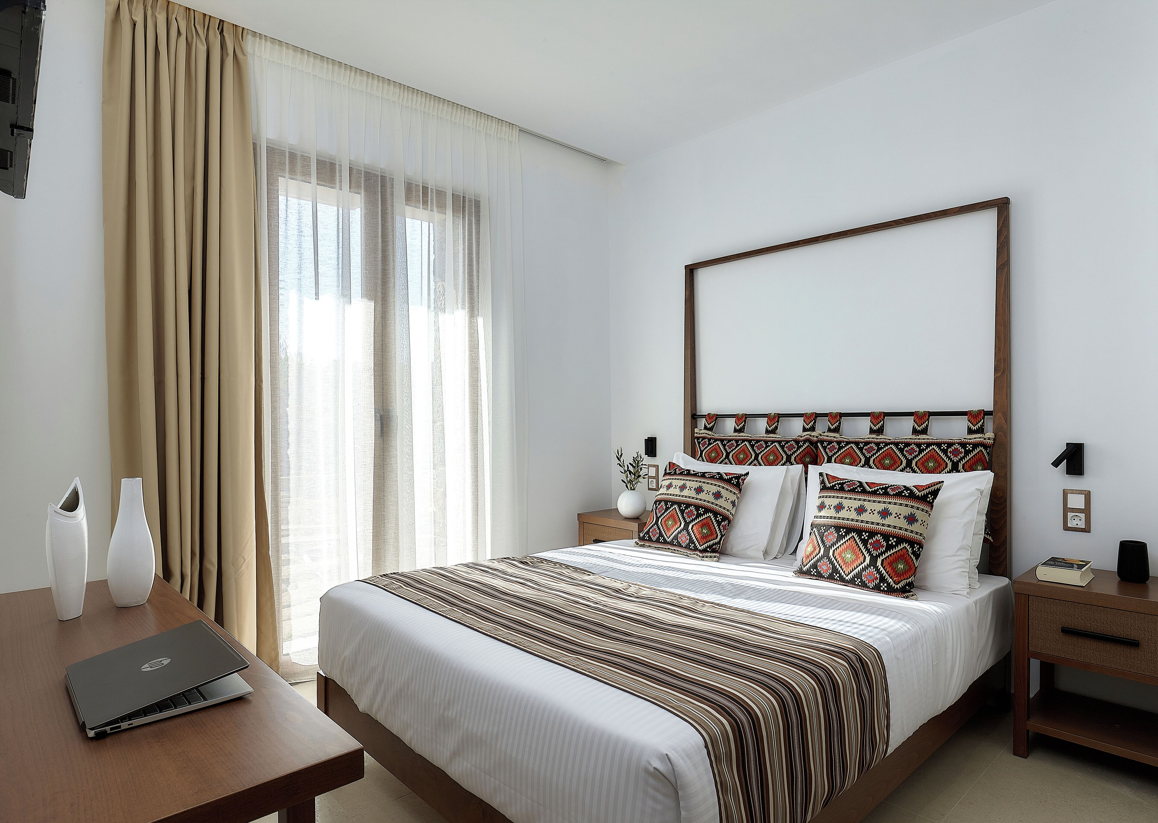 A hotel room with a large bed, a wooden headboard, a bedside table with a laptop, and a window with curtains.