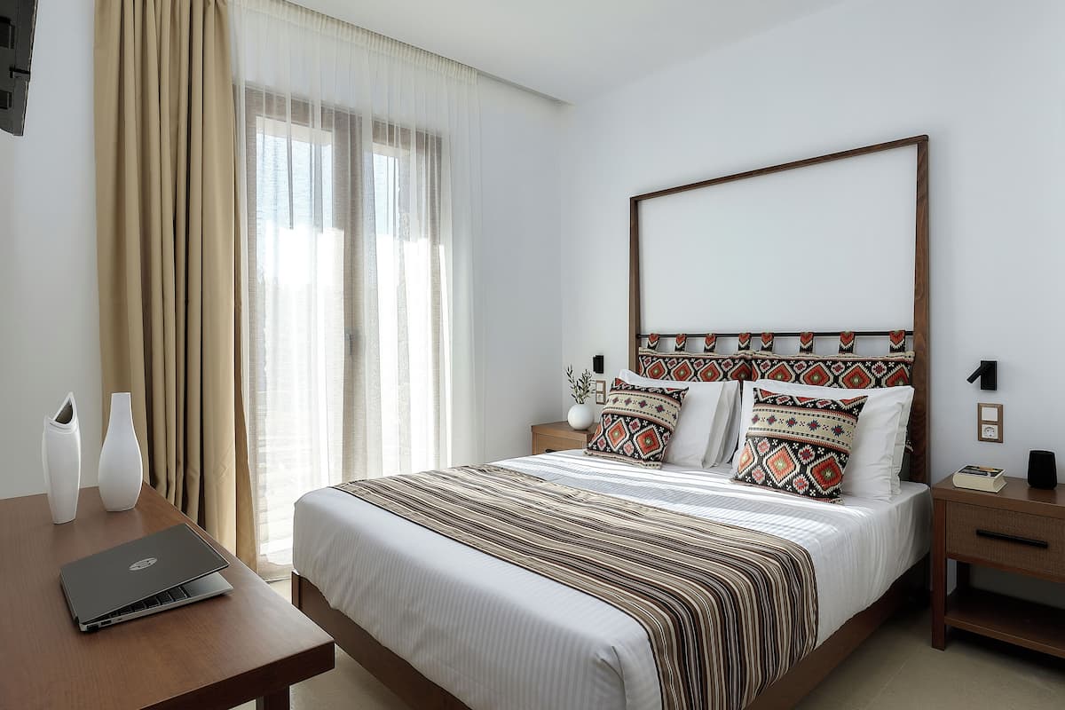 Ein Hotelzimmer mit einem großen Bett, einem hölzernen Kopfteil, einem Nachttisch mit einem Laptop und einem Fenster mit Vorhängen.