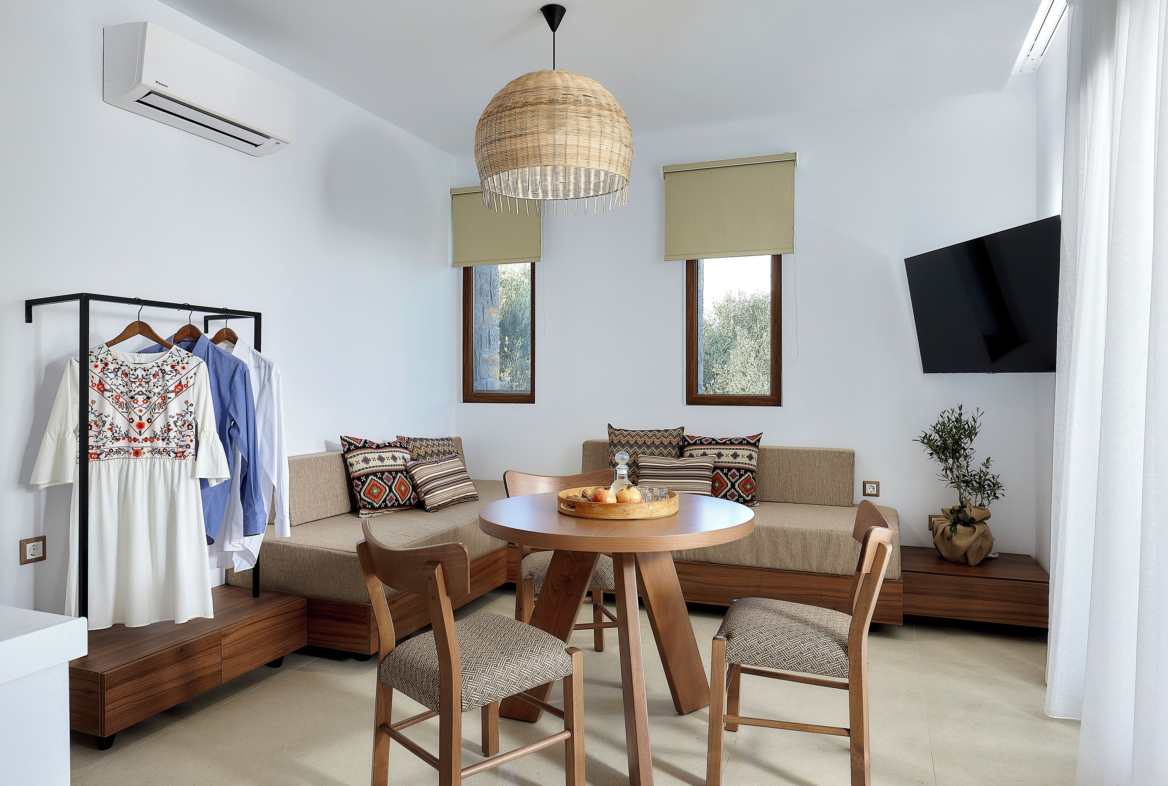 Un salon moderne avec une table ronde en bois, deux canapés, un téléviseur fixé au mur et un portant à vêtements avec des vêtements suspendus.