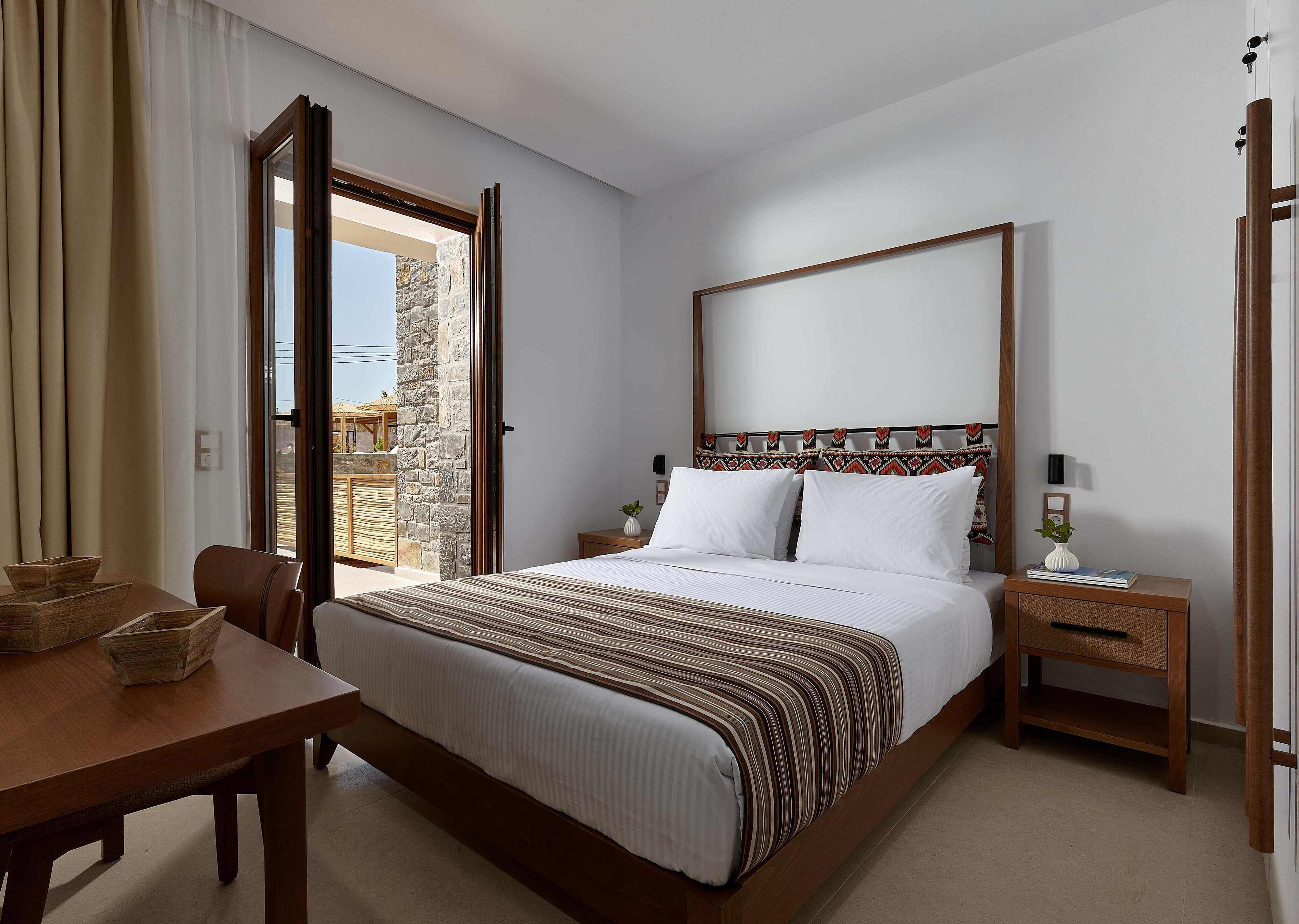 Quarto de hotel com cama, mesa de cabeceira de madeira, mesa com cadeiras e vista através de uma porta de vidro.