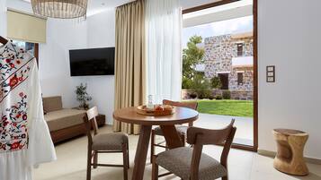 Ein modernes Wohnzimmer mit einer Couch, einem Esstisch mit Stühlen, einem an der Wand befestigten Fernseher und Blick auf ein Steinhaus durch ein großes Fenster.