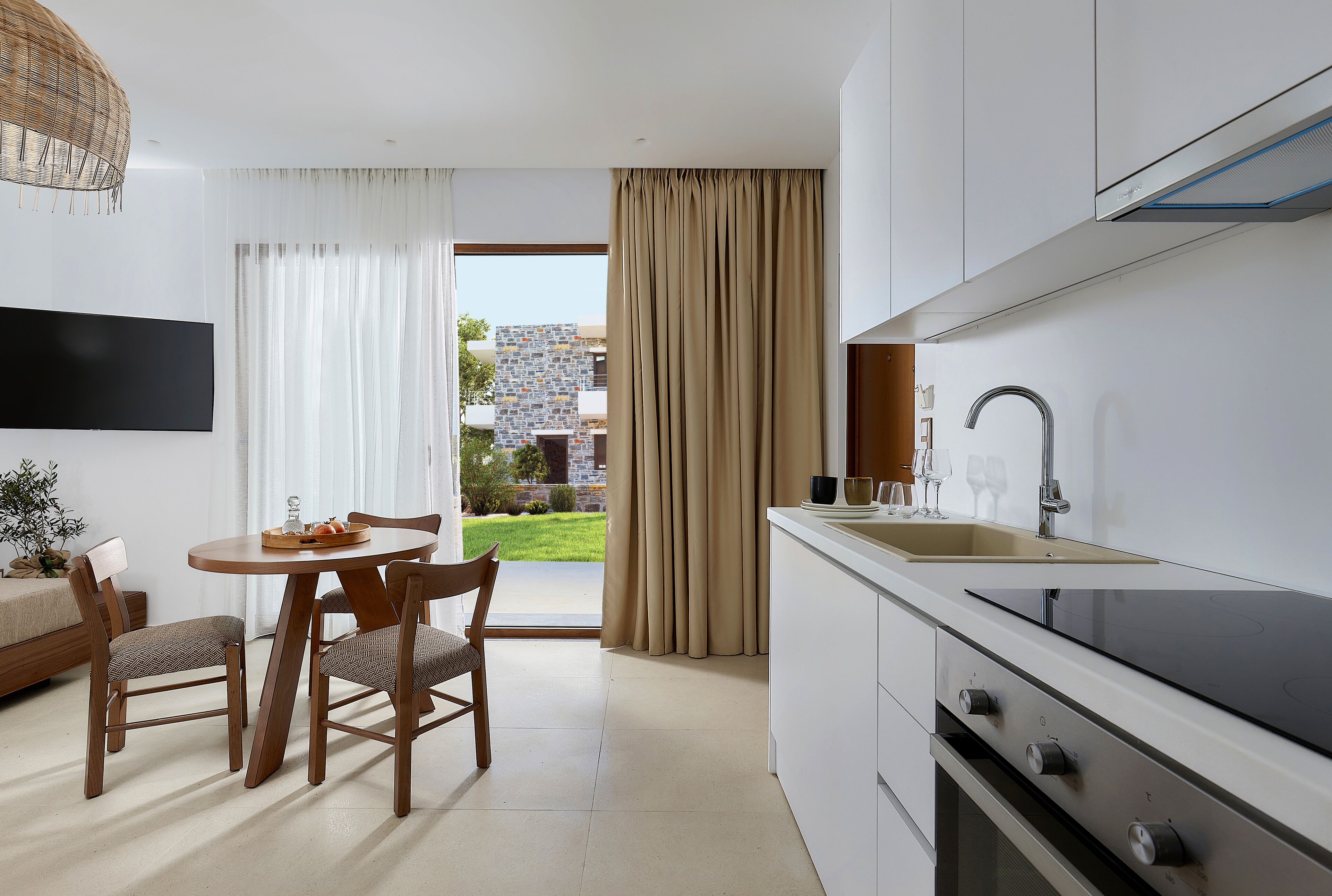 A modern kitchen with a built-in stove, a dining area with a round table and chairs, and a view of a garden through large windows.