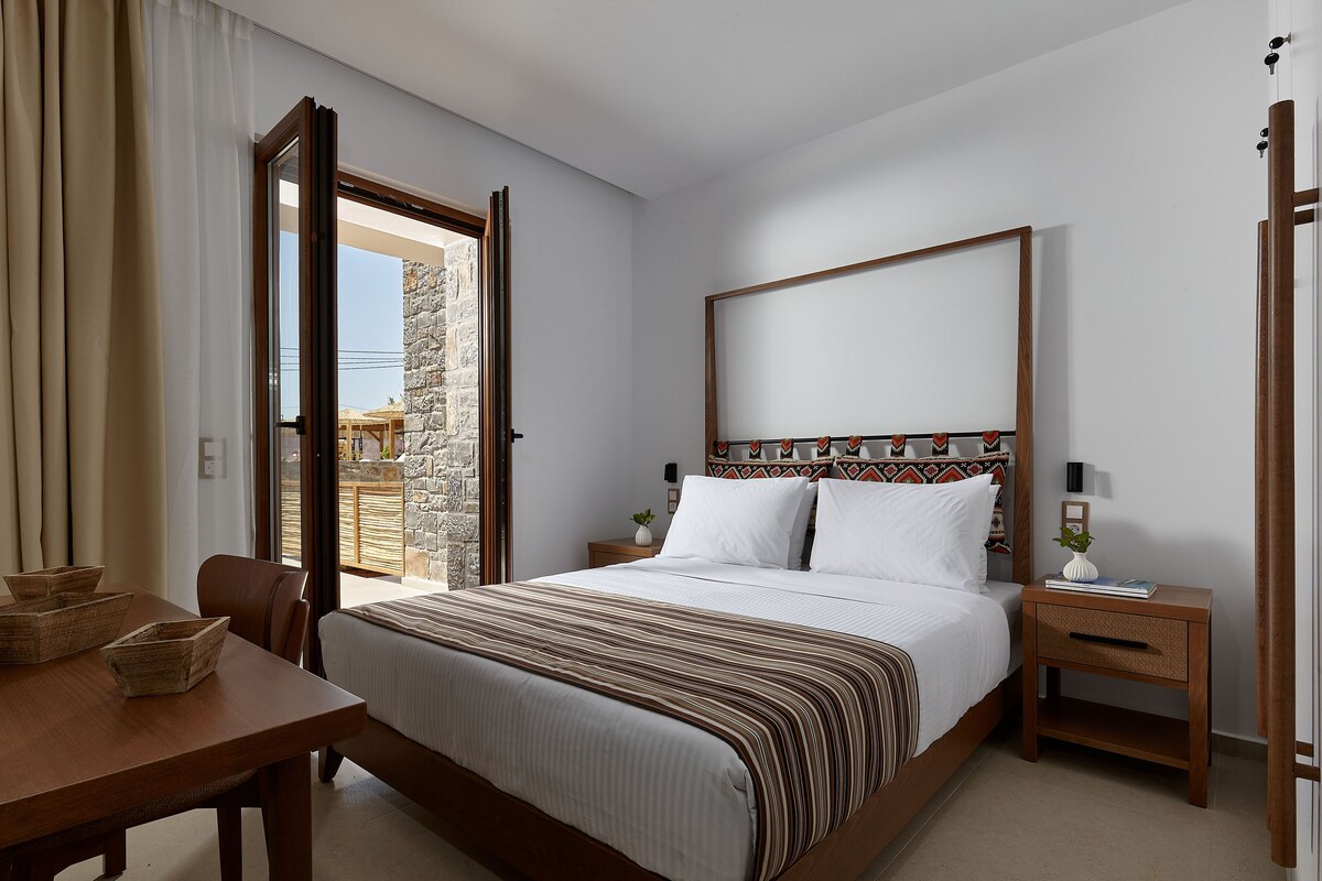 Ein Schlafzimmer mit einem Bett, Nachttischen, einem Holznachtkästchen, einem Tisch mit Stühlen und Blick ins Freie.