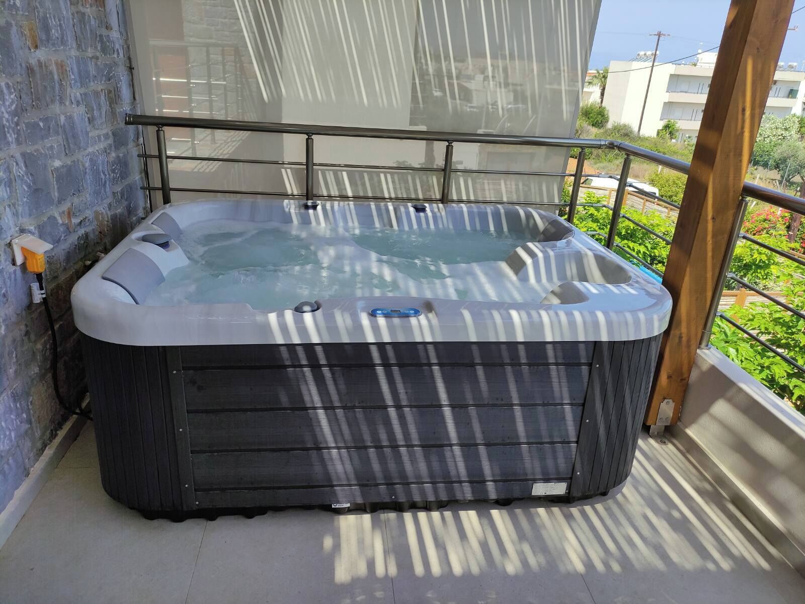 Un jacuzzi sur un balcon doté d’une rambarde en métal, d’un mur en briques et offrant une vue sur des bâtiments et de la verdure.