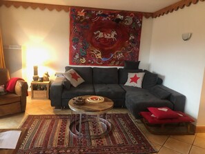A living room with a sofa, a coffee table, a patterned rug, a tapestry, and a small side table with decorative items. - Charming Chalet-Style Apartment with Terrace and Peaceful Setting, Close to Slopes and Amenities (Les Menuires)