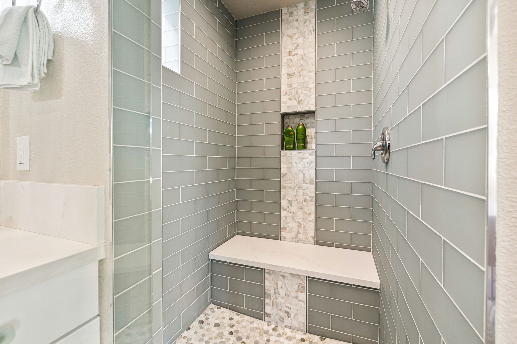 A shower area with grey tiles, a bench, and two green bottles on a shelf.