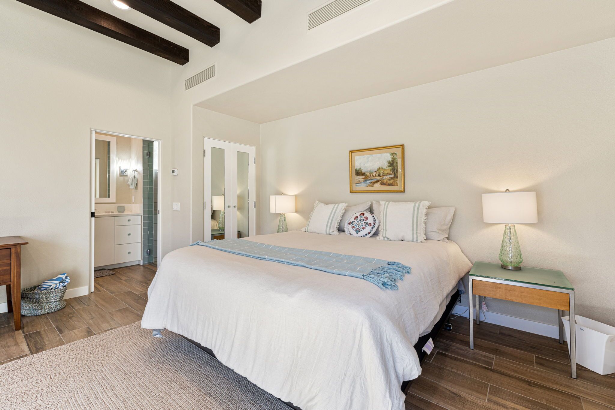 A bedroom with a large bed, bedside lamps, a painting on the wall, and a small table with a lamp.