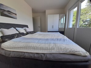 Une chambre à coucher moderne avec un grand lit, une tête de lit, une armoire et une fenêtre donnant sur des arbres.