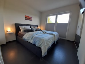 Une chambre à coucher comprenant un lit, des tables de chevet, une commode, un tableau et une porte-fenêtre coulissante donnant sur l’extérieur.