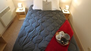 A single bed with a blue quilt, white pillows, and a red throw blanket. There are bedside tables with lamps and a tray with cups and a spoon.