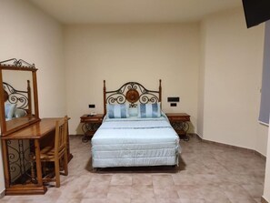 Un dormitorio con una cama, una mesita de noche de madera, un tocador, un espejo y un televisor instalado en la pared.