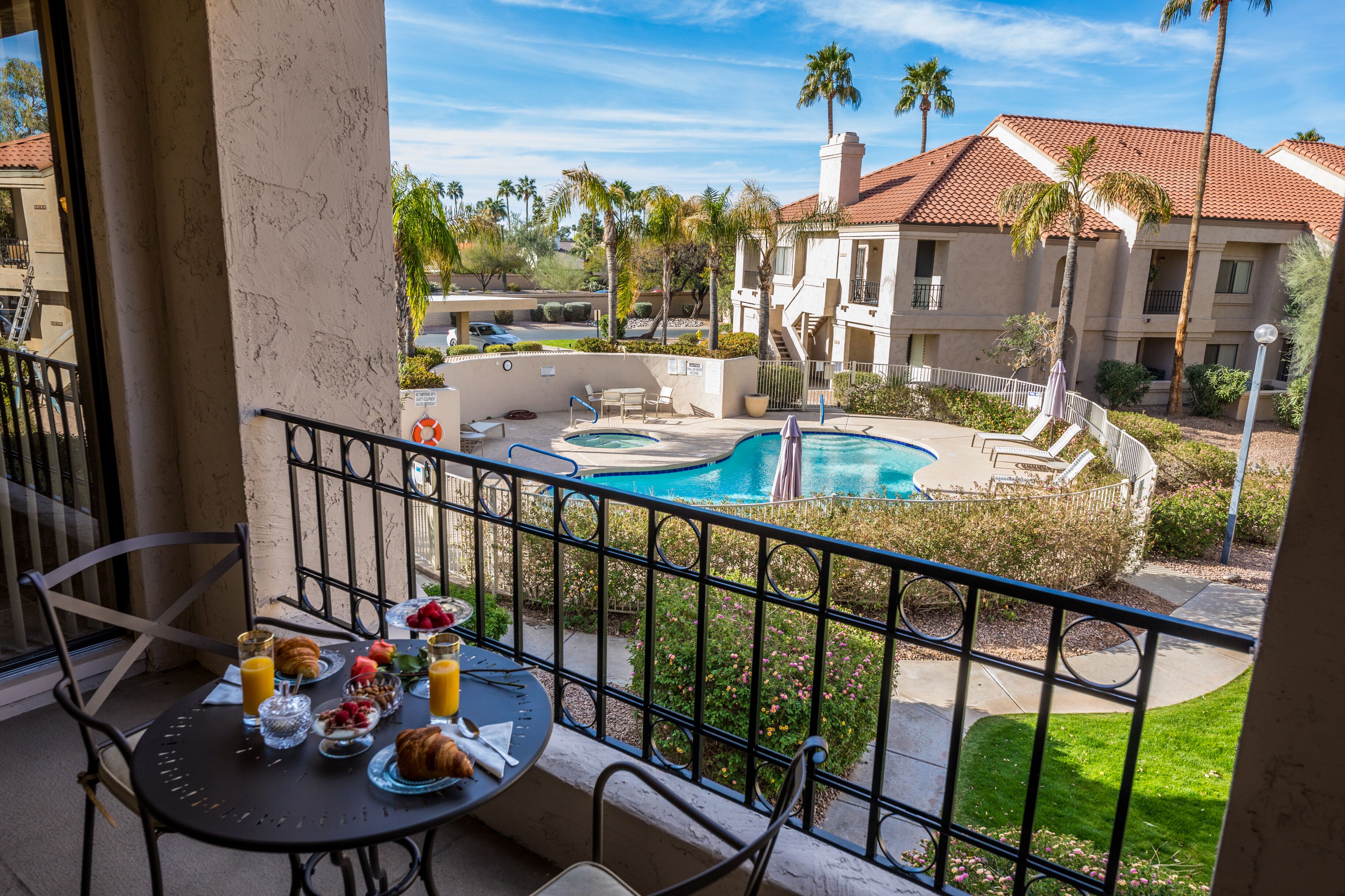 A balcony with a table set for breakfast, overlooking a swimming pool and a building with a red-tiled roof.