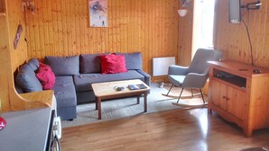 Una sala de estar acogedora con un revestimiento de madera, un sofá gris con cojines rojos, una mesa de centro de madera, un sillón mecedor y un armario de madera.