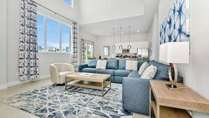A spacious living room with a blue sofa, a white armchair, and a coffee table. A large window with curtains is visible, and a kitchen area is in the background.
