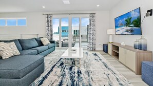 Una sala de estar moderna con sofá, mesa de centro y televisor. Hay un balcón con vista al mar y edificios.