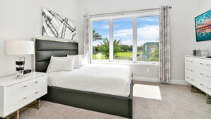 A modern bedroom with a large bed, a nightstand, a dresser, and a window with a view of greenery.
