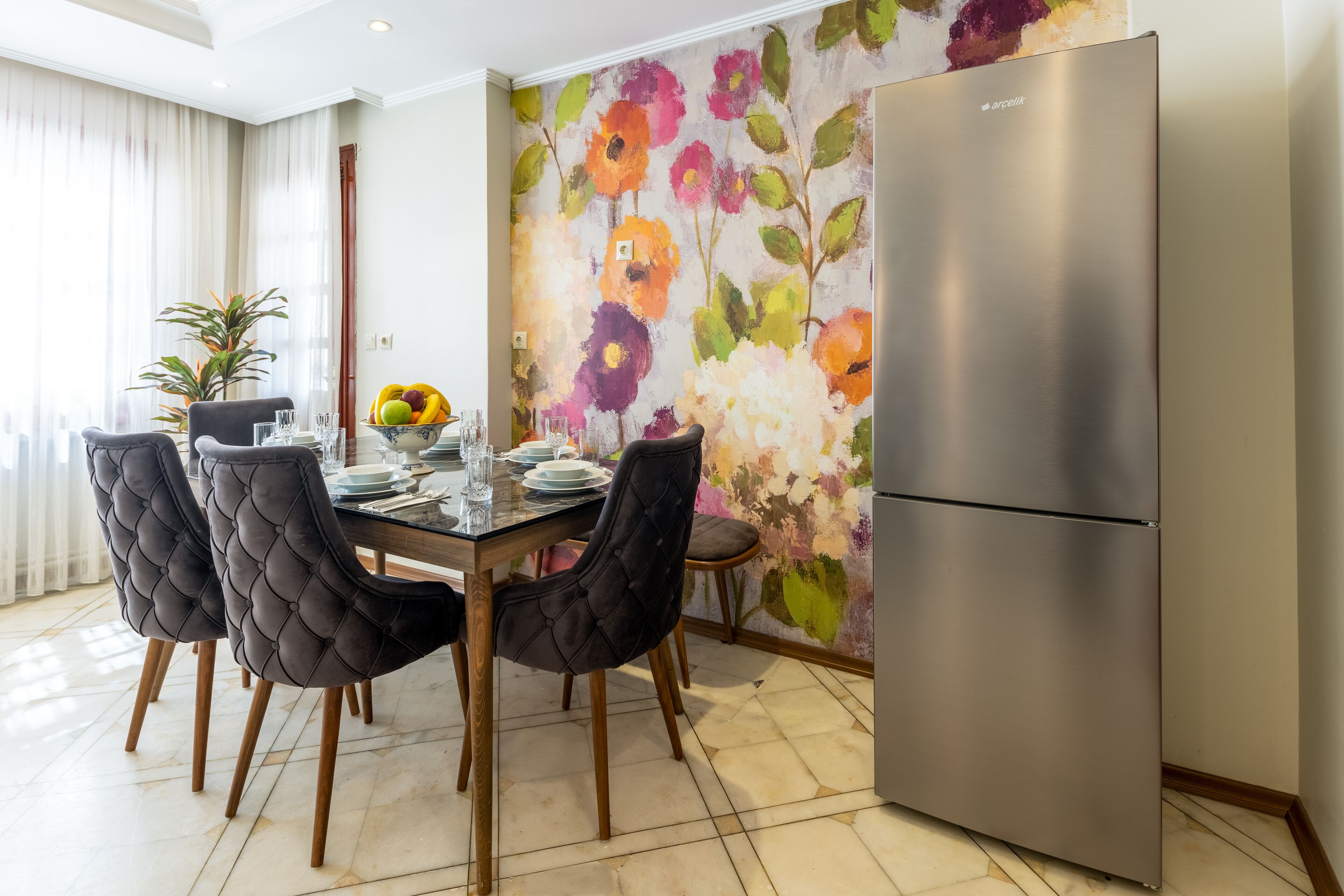 Un comedor con una mesa dispuesta para cuatro, rodeada de sillas capitonadas, un refrigerador moderno y un mural floral llamativo.