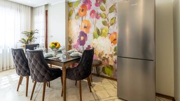 Un comedor con una mesa dispuesta para cuatro, rodeada de sillas capitonadas, un refrigerador moderno y un mural floral llamativo.