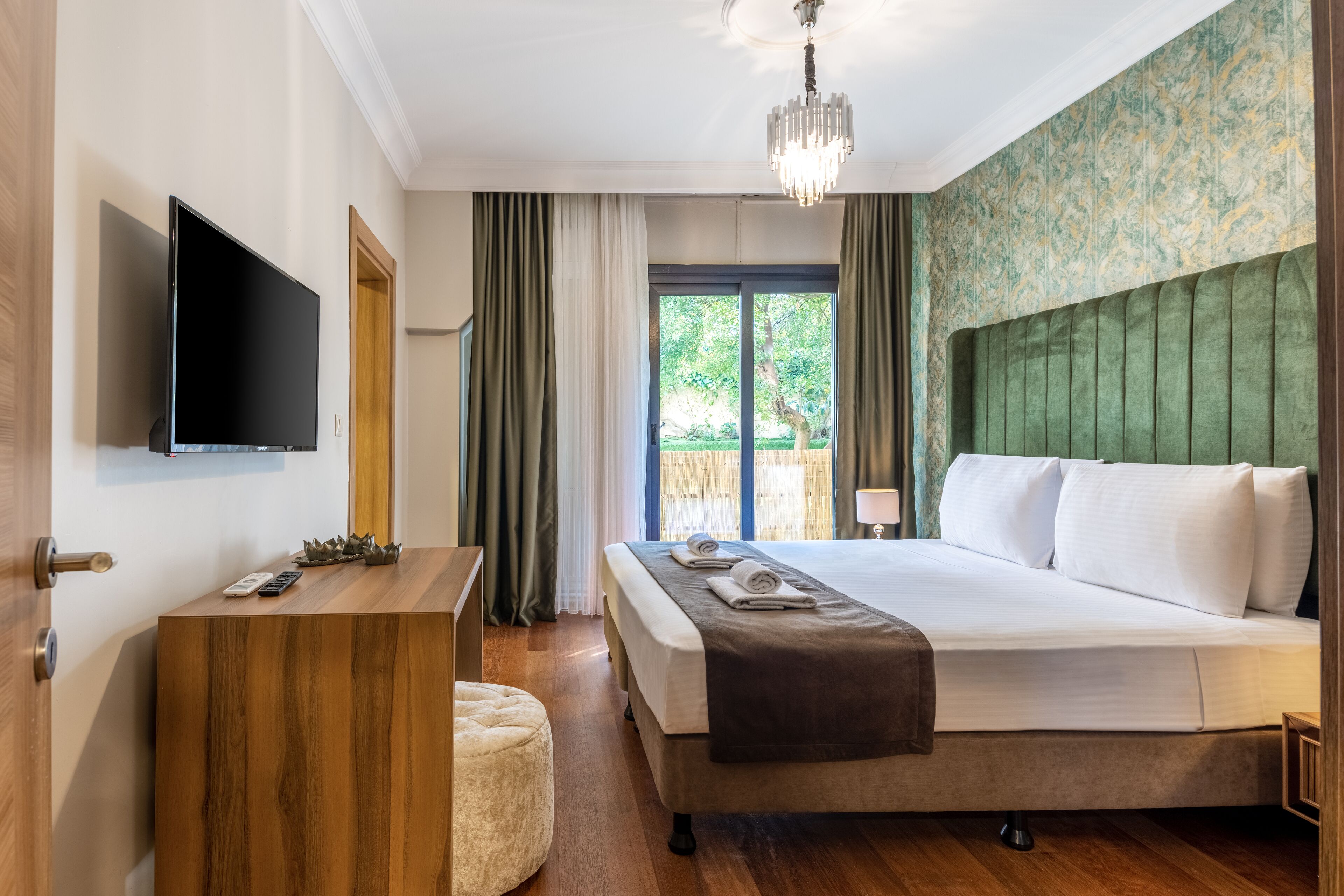 A hotel room with a bed, a desk, a TV, and a view of greenery outside.