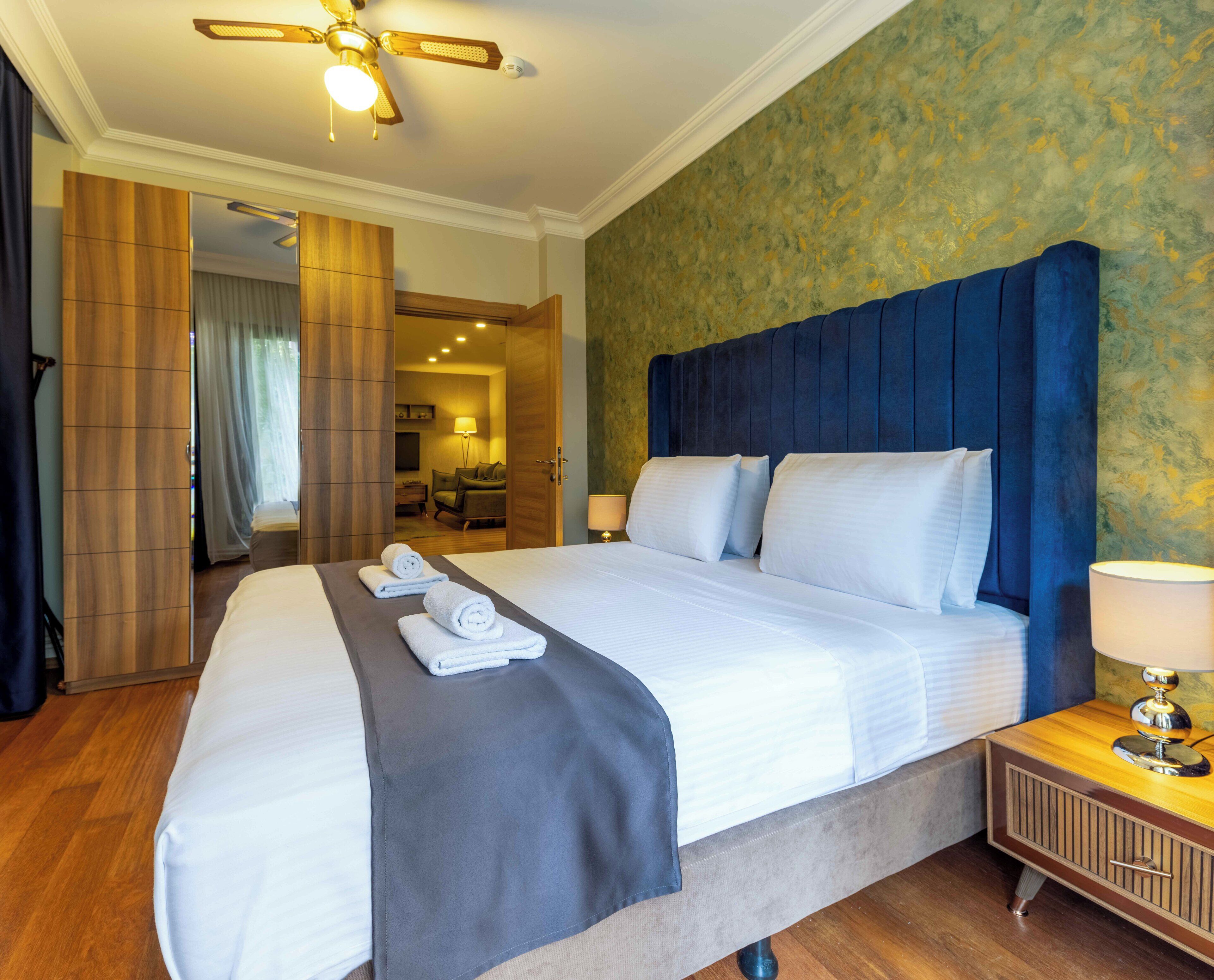 A hotel room with a large bed, a wooden wardrobe, a bedside table with a lamp, and a ceiling fan.