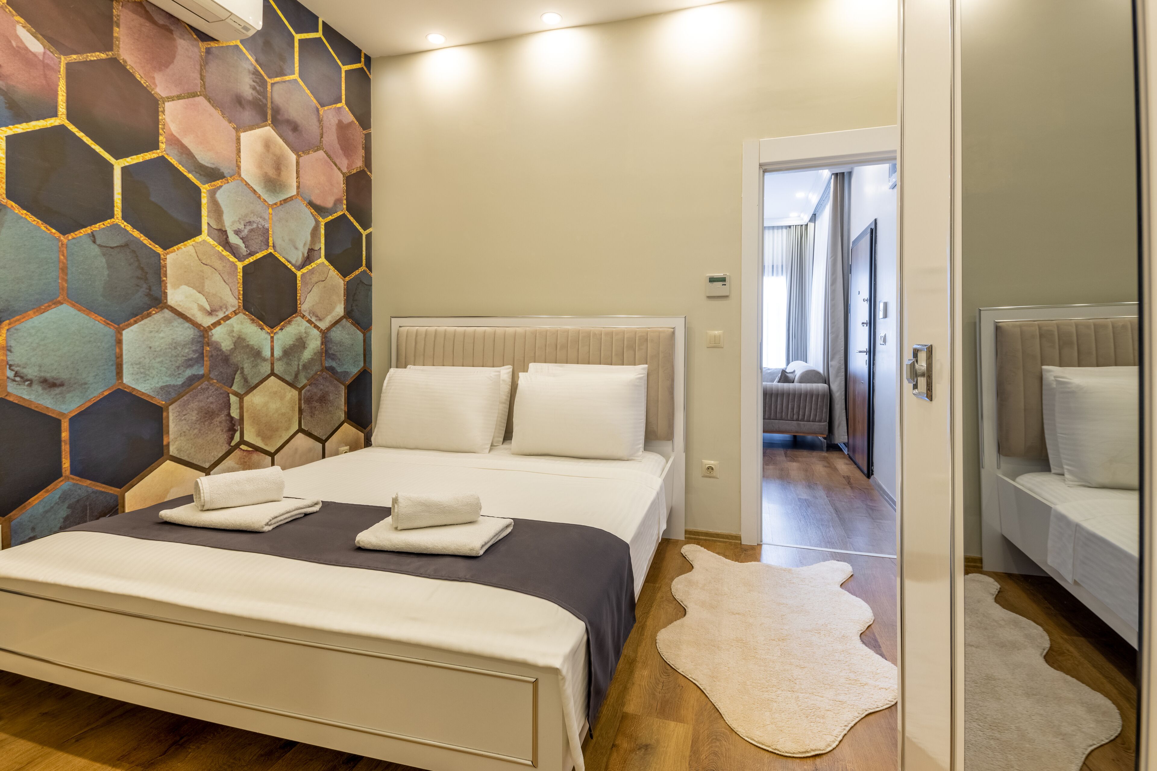 A modern hotel room with a large bed, a unique hexagonal wall design, and a separate bedroom visible through an open door.