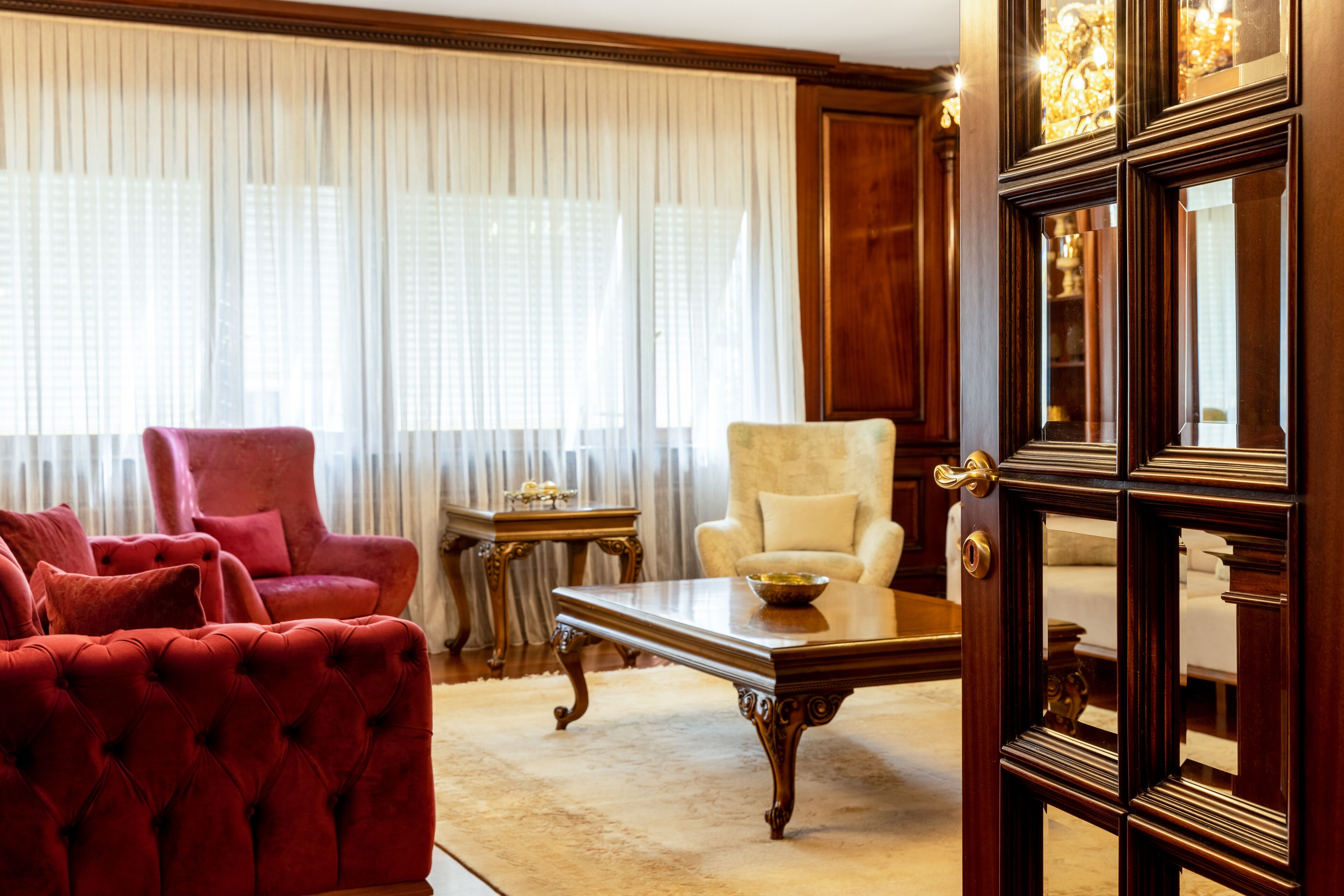 A room with a red tufted sofa, a cream armchair, a wooden coffee table, and a large mirror.