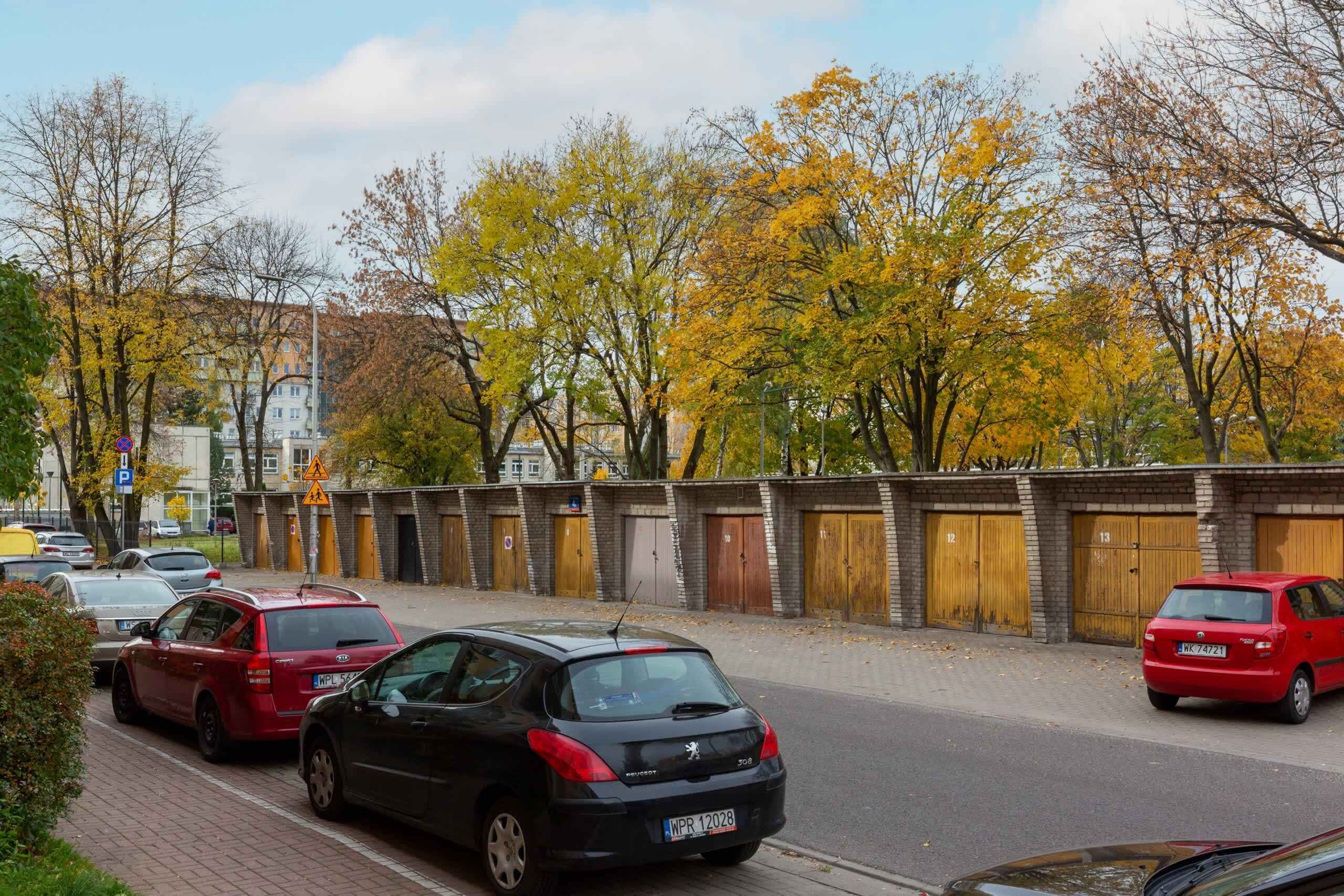 Une rue bordée de voitures garées, un mur de briques avec des portes numérotées et des arbres aux feuilles d’automne.