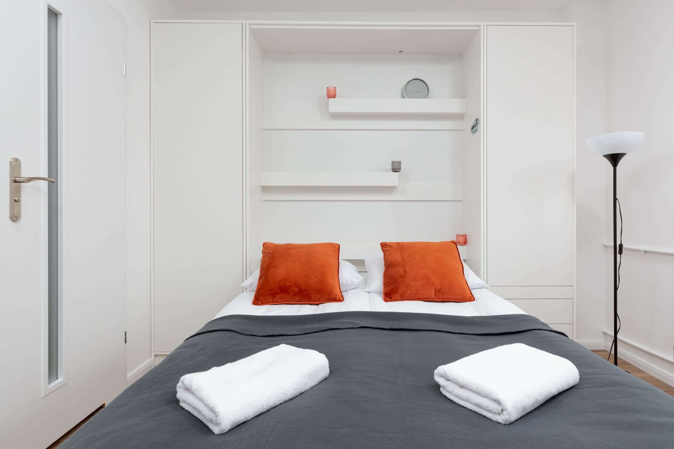 Ett hotellrum med en säng, två orange kuddar, vita handdukar, en sänglampa och en vit garderob med hyllor.