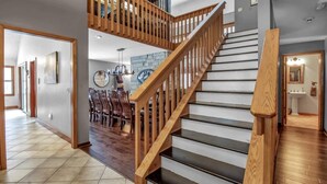 Un interior espacioso con una escalera de madera que conduce a un segundo piso, un comedor con sillas y una cocina visible al fondo.
