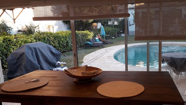 A wooden table with a woven basket and two woven placemats, a covered grill, and a view of a swimming pool and outdoor seating area through large windows.