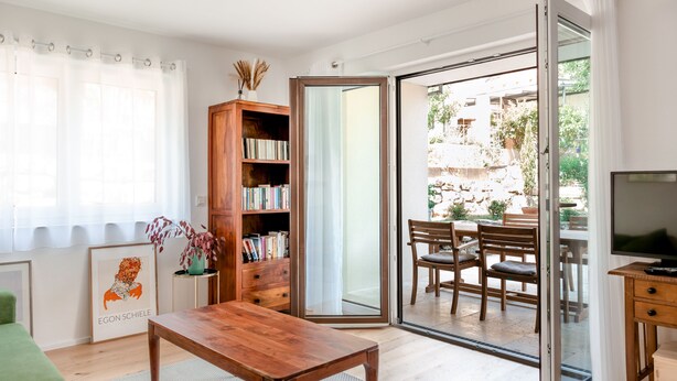 A living room with a wooden table, a bookshelf, a framed picture, and a sliding glass door leading to an outdoor patio with a table and chairs. - Apartment `Akazie' in Radolfzell-Liggeringen (Radolfzell am Bodensee)