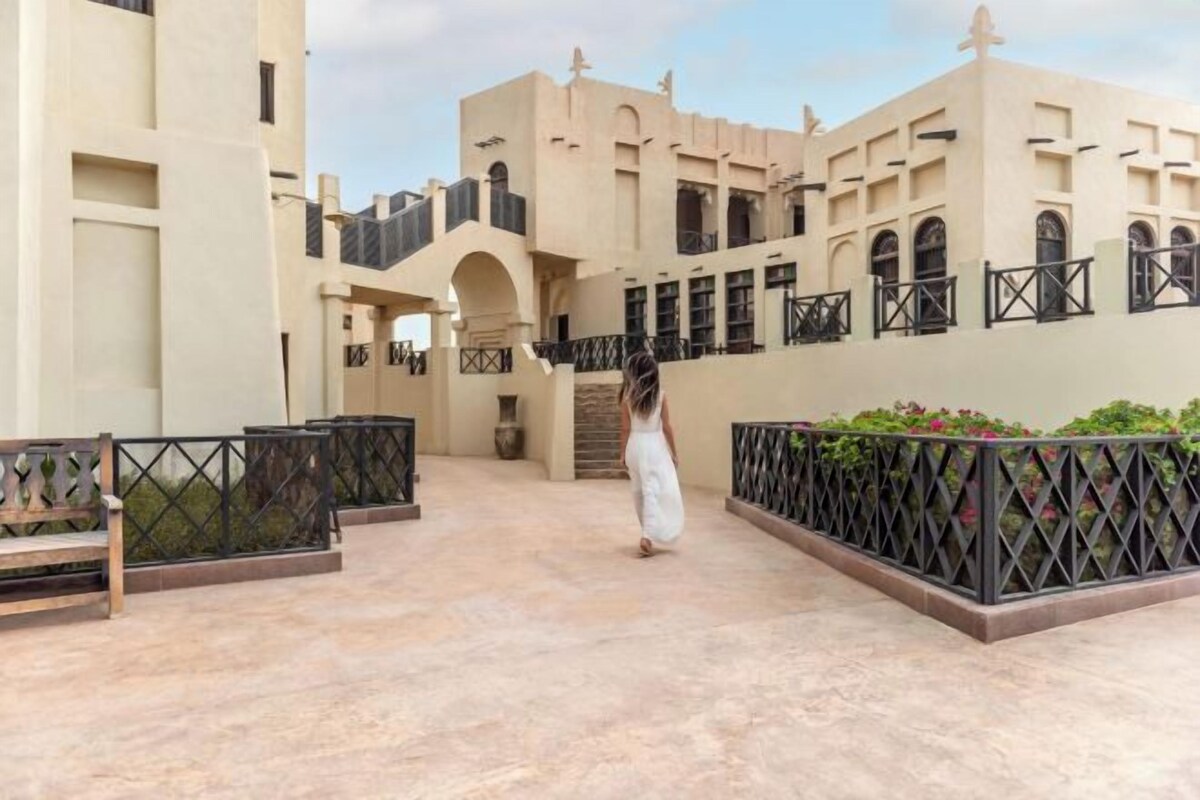 A courtyard with a person walking, beige buildings with arches, and a staircase.