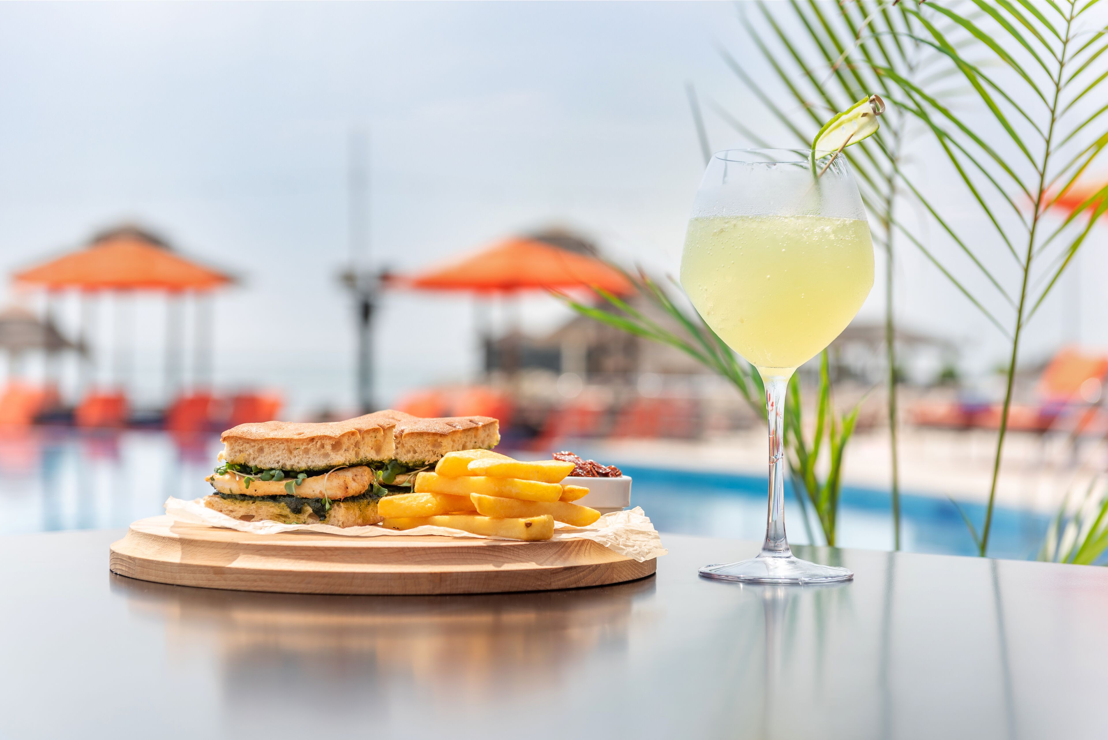 A meal with a sandwich and fries on a wooden board, a glass of a yellow beverage, and a poolside setting with umbrellas in the background.