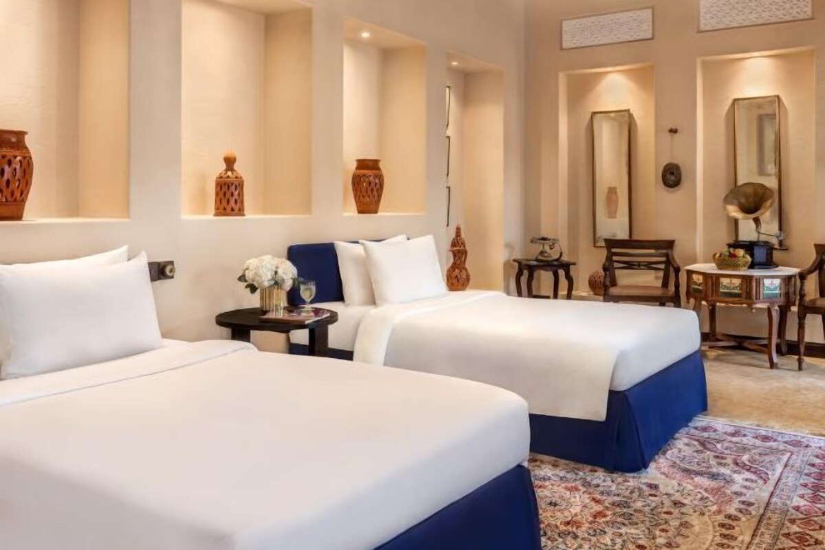 A hotel room with two beds, a small table with a vase, a chair, and a mirror on the wall.