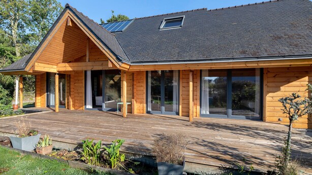 Una moderna casa in legno con tetto spiovente, ampie finestre e un terrazza in legno.