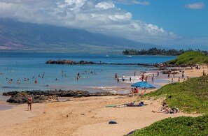 Ein Strand mit badenden und sonnenden Personen, einem Felsen und einem entfernten Gebirgszug.