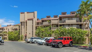 A parking lot with several vehicles, including a red Jeep, and a building with multiple balconies and palm trees.
