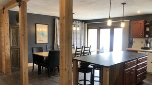 A kitchen with wooden beams, a dining area with a large table and chairs, and a view of the outside through a window.
