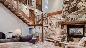 A spacious living area with a high ceiling, a stone fireplace, a wooden staircase, and a comfortable seating arrangement. - Moose on the Loose: Private Hot Tub, Mountain Views, Quick Ski Access (Breckenridge)