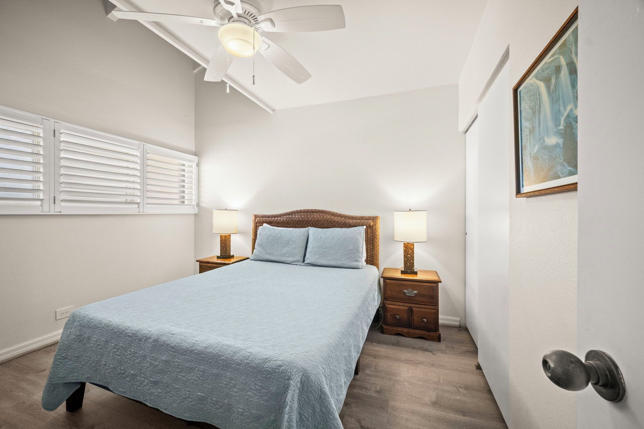A bedroom with a bed, two bedside tables with lamps, a ceiling fan, and a painting on the wall.