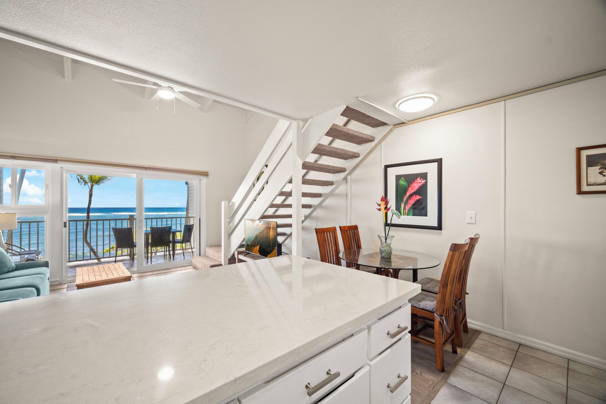 A modern kitchen with a large island, a dining area with wooden chairs, a staircase, and a balcony with a view of the ocean.
