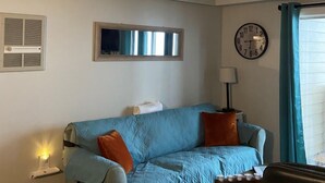 Un salon avec un canapé bleu clair, une horloge, une fenêtre avec des rideaux et un radiateur fixé au mur.