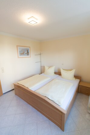 A bedroom with a wooden bed, white bedding, a painting on the wall, and a ceiling light fixture.