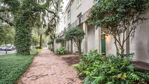 A brick pathway lined with greenery and trees, leading to a building with a green door.