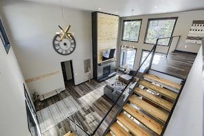 A modern living room with a fireplace, large windows, and a staircase with wooden railings.