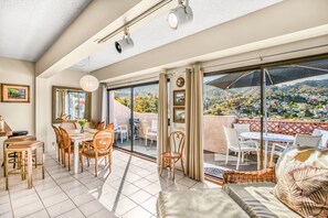 A spacious living area with a balcony, furnished with wicker chairs and a dining table, overlooking a scenic hillside.