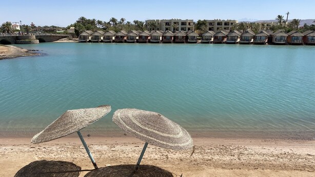 Een strand met twee rieten strandparasols en een rij bungalows boven het water op de achtergrond.